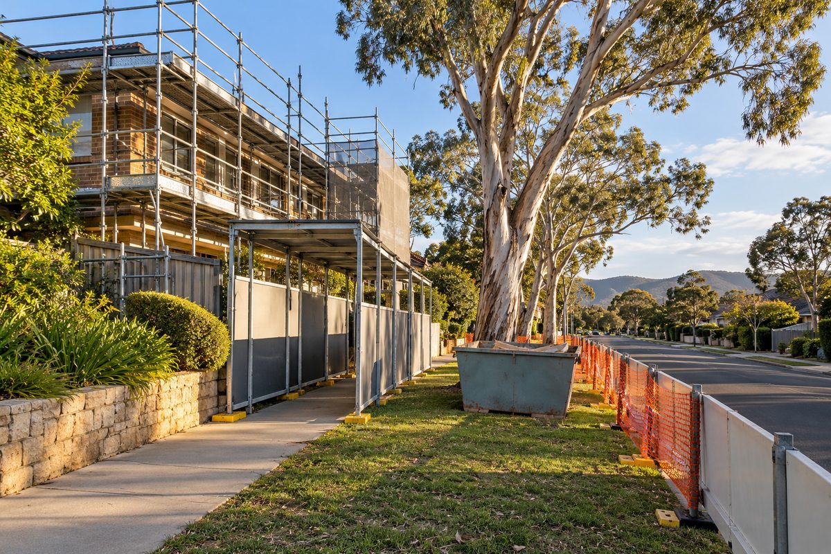 A Canberra street with a scaffold on a house, a short covered walkway and barrier panels keeping the footpath clear, orange mesh fencing at the kerb and a skip bin on the nature strip beneath ghost gums